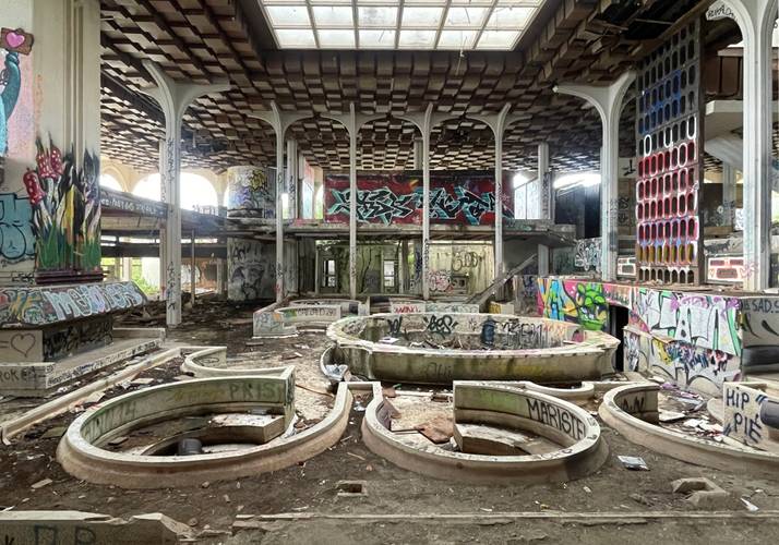 Verlassener Raum mit verzierten Säulen, bunten Graffiti an Wänden und Decke, mehrere leere Brunnen. Boden voller Schutt und Abfall.