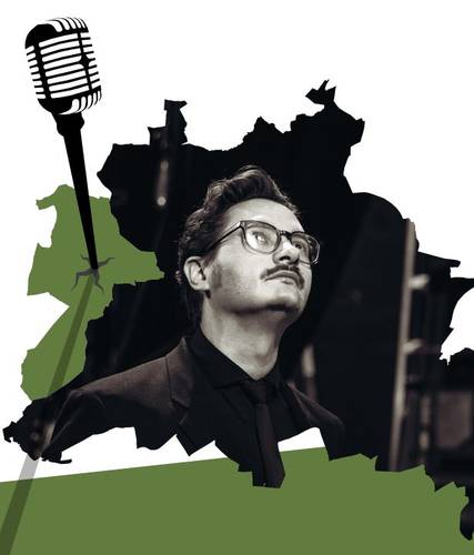 Männlich-gelesene Person mit Brille, Anzug und Schnurrbart, im Hintergrund ein schwarzes Mikrofon und grüne Karte.