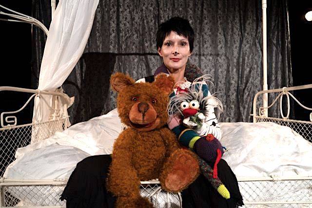 Person mit kurzem dunklem Haar auf einem Bett hält Grossteddybaer und bunte Handpuppe mit weissen Haaren vor einem dunklen Vorhang.