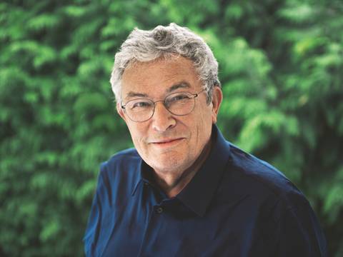 Charles Lewinsky – Ältere Person mit Brille und blauem Hemd vor grünem, unscharfem Hintergrund mit Blättern.