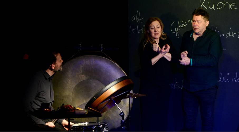 Drei Personen, eine mit Drum und Gong, zwei gestikulierend vor Tafel mit den Worten alte, Tele, Geburt, Küche.