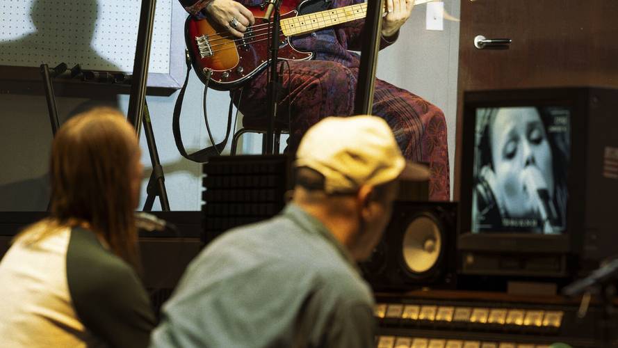 Stereophonic – Person spielt Bassgitarre in Kabine. Zwei Personen mit Baseballkappe und langen Haaren beobachten im Studio. Monitor zeigt Nahaufnahme einer Sängerin.