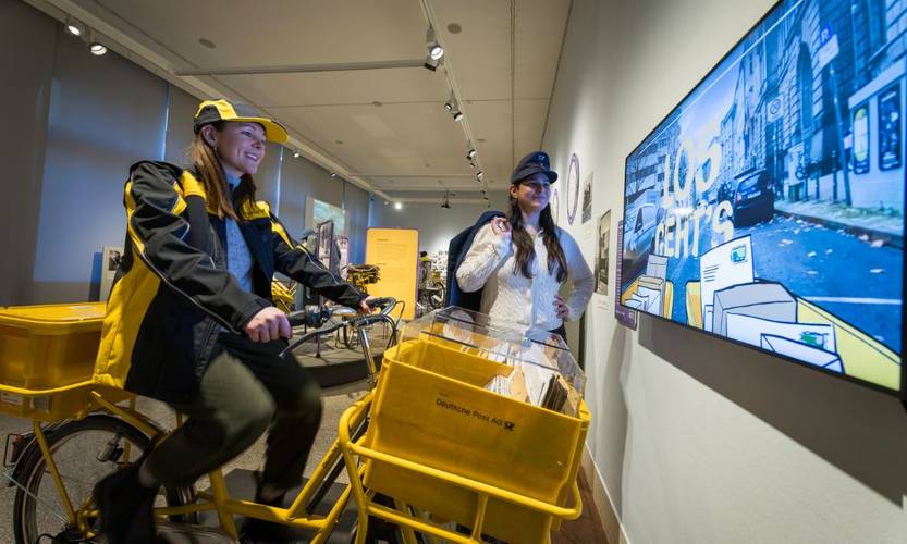 Blick in die Ausstellung "niemals radlos" – Eine Frau auf einem gelben Postfahrrad in der Ausstellung "niemals radlos"