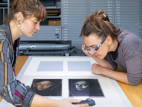 Die Restauratorinnen Tabea Harney und Antje Penz untersuchen zwei Lithographien von Käthe Kollwitz. – Zwei Personen betrachten Lithographien auf einem Tisch, eine mit Stirnlampe. Beide tragen lässige Kleidung. Die Drucke zeigen ein Porträt eines Gesichts von Käthe Kollwitz.