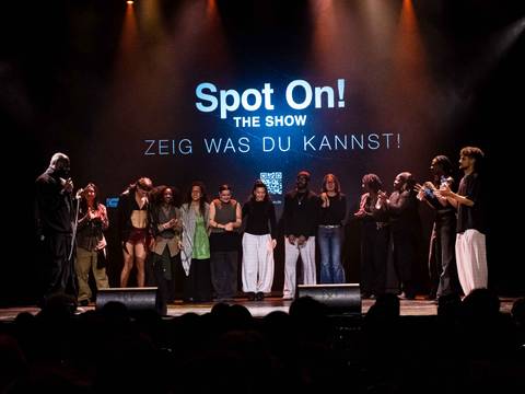  – Gruppe von 11 Personen auf Buehne, einige klatschen, andere schauen zur Seite. Hintergrundtext: "Spot On! The Show ZEIG WAS DU KANNST!".