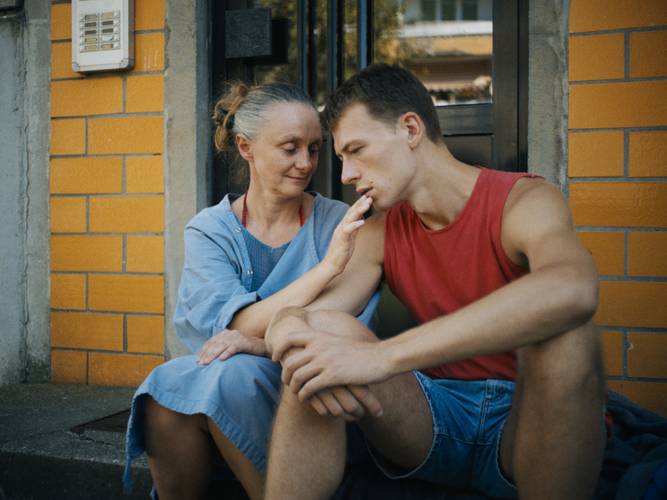 Man sieht einen jungen Mann und eine ältere Frau auf einem Treppenabsatz sitzen. Die Frau hält ihre Hand an den Mund des Mannes. © Grandfilm – Man sieht einen jungen Mann und eine ältere Frau auf einem Treppenabsatz sitzen. Die Frau hält ihre Hand an den Mund des Mannes.