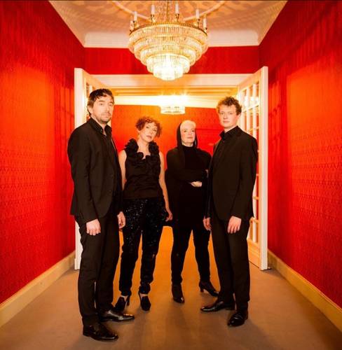 Vier Personen in schwarzer Abendgarderobe stehen in einem elegant beleuchteten Flur mit leuchtend roten Wänden, weißen Türrahmen und zwei großen Kristalllüstern. Die Gruppe blickt in die Kamera und ist mittig im Bild positioniert. Die warme Beleuchtung und die symmetrische Perspektive verleihen der Szene eine festliche, stilvolle Atmosphäre.