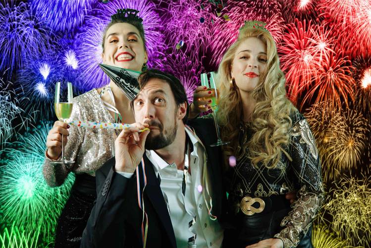 Chaos Royal – Drei Personen mit Sektglaesern feiern vor buntem Feuerwerk. Eine Person traegt einen Partyhut, eine bläst in einen Papiertröte.