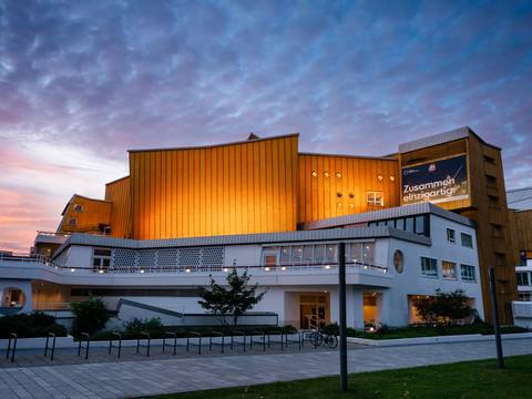 Die Philharmonie im Abendlicht – Die Philharmonie Berlin mit goldenen Panelen und weißen unteren Stockwerken wird in der Abenddämmerung beleuchtet, mit einem bewölkten Himmel im Hintergrund. Auf einem großen Banner steht auf Deutsch „Zusammen einzigartig“.