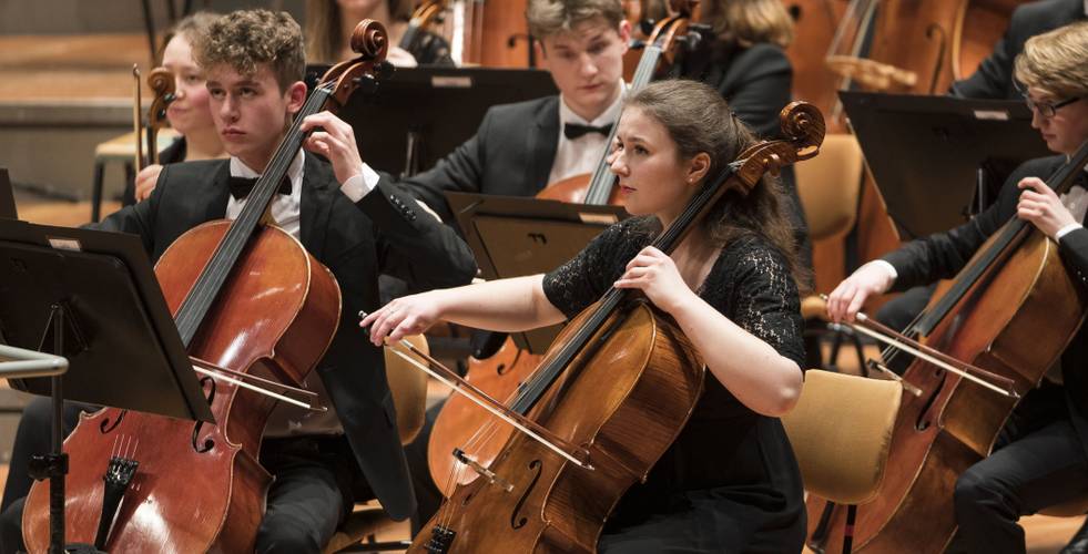 Personen mit Celli in einem Orchester, formelle Kleidung, konzentriert spielend, Notenstaender im Hintergrund.