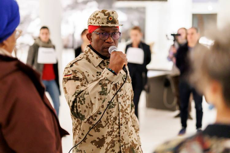 David Dibiah – Person in sandfarbener Militär-Uniform mit Mikrofon spricht, andere Personen im Hintergrund, unscharf.