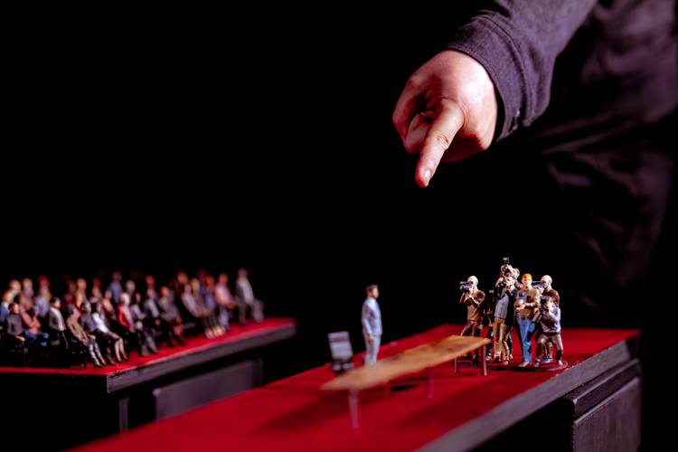 Transport: Tipping Point – Große Hand zeigt auf Miniaturfiguren auf einem roten Laufsteg: Fotograf*innen umringen eine stehende Person, hinten sitzt Publikum.