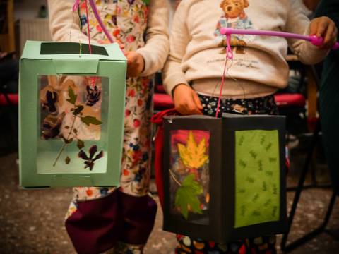  – Zwei Kinder halten bunte Papierlaternen mit gepressten Blättern und Blüten. Eine Laterne ist grün, die andere schwarz mit grünem Dekor.