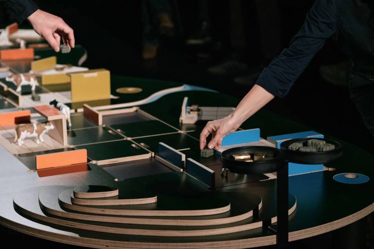 The Game – Zwei Hände positionieren grüne Objekte auf einem Architekturmodell mit bunten Strukturen und kleinen Tierfiguren auf schwarzem Untergrund.
