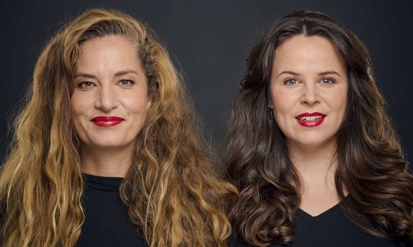 Suchtpotenzial – Zwei Personen mit langen Haaren und roten Lippenstift, beide tragen schwarze Oberteile, blicken frontal in die Kamera vor grauem Hintergrund.