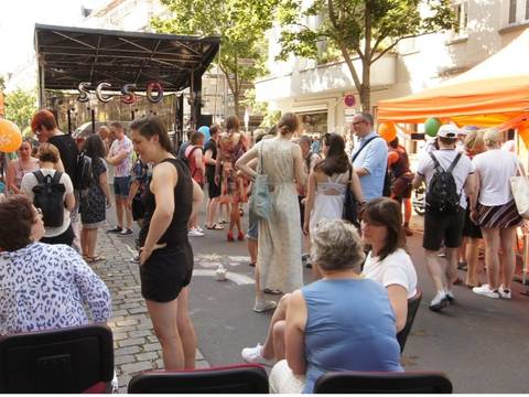  – Personengruppe bei Straßenfest mit orangefarbenen Zelten, Luftballons und Informationsstand. Im Hintergrund Bühne mit Schriftzug "SCSO". Rechts Flagge von