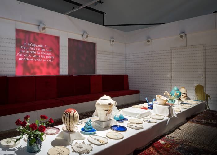 Rote Sitzbänke an der Wand, darüber rotes Licht mit Text. Langer Tisch mit Kunstobjekten wie Vasen und Tellern, rote Rosen im Vordergrund.