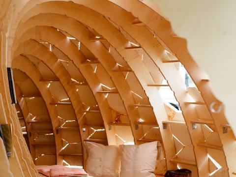 MY-CO-X, MY-CO Space, 2021 – Foto: Blick in einen höhlenförmigen, halbrunden Bau aus Holz mit Sitzbank, einem Teppich und Kissen. Von außen strömt durch Spalten Tageslicht nach innen.