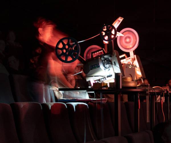 Warm Data – Filmprojektor mit rotierenden Filmspulen in einem dunklen Raum auf klappbaren Kinositzen. Verschwommenes Abbild einer Person daneben.