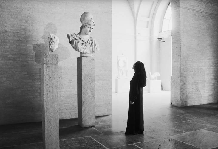 Arbeit am Mythos. Elshan Ghasimi vor der Athena in der Münchner Glyptothek, 2024 Foto: Georg Stirnweiß – Schwarzweißfoto einer Person in langer Kleidung vor antiken Büsten auf Säulen in einem Museumsraum mit gemauerter Wand und großen Fenstern.