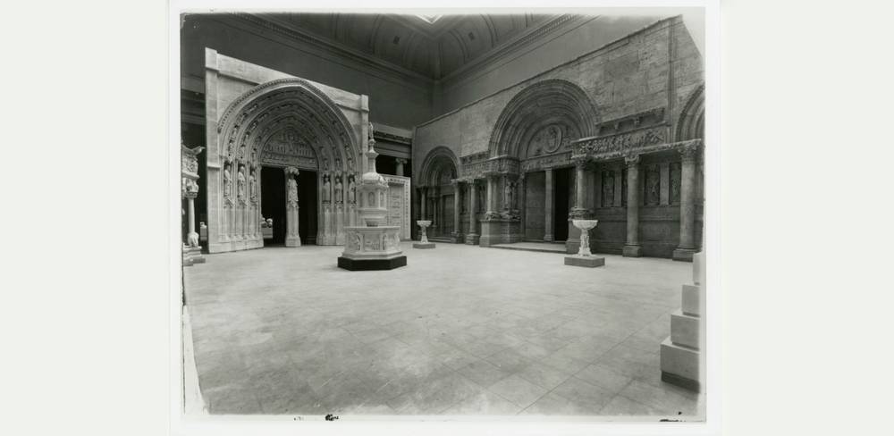 Ein Abguss des Taufbeckens in der Architectural Hall des Carnegie Museum of Art, Pittsburgh, nach 1908 – Schwarzweiß-Fotografie eines Museumsraums mit romanischen Bögen und Arkaden. Große steinerne Verzierungen und ein freistehendes, reich verziertes Taufbecken.