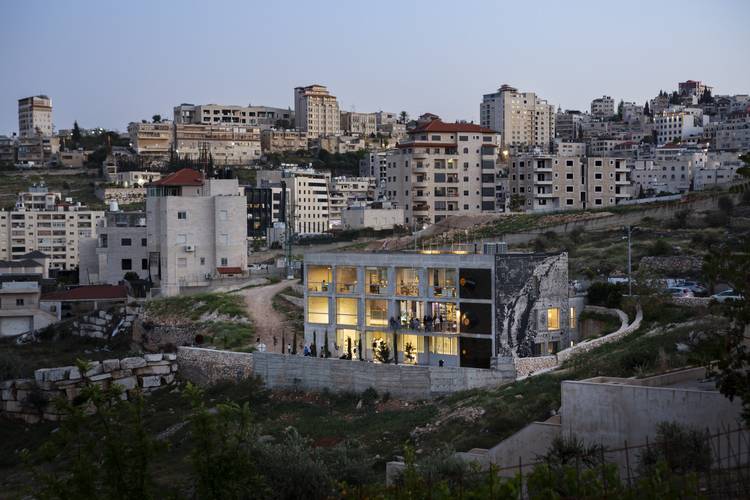 AAU Anastas, The Wonder Cabinet / Cultural Center, Bethlehem, 2023 Foto: Mikaela Burstow – Ein modernes, zweistöckiges Gebäude mit Glasfront erleuchtet in urbaner Umgebung; im Hintergrund dichte Wohnbebauung auf Hügeln.