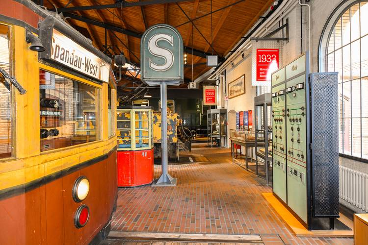 Der Führerstand eines Berliner S-Bahn-Triebwagens von 1927 erinnert an die Einführung der Stadt-, Ring- und Vorortbahnen nach dem ersten Weltkrieg. Sie werden bis heute über eine seitliche Stromschiene versorgt, die speziell hierfür entwickelt wurde – Der rot-gelbe Führerstand eines S-Bahnwagens steht im Vordergrund, dahinter steht ein großes S-Bahn Schild in der Ausstellung.