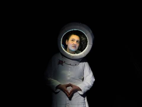  – Person in Astronautenkostüm im Dunkeln, beleuchtetes Gesicht im Helm, Hände vor dem Körper gefaltet, nachdenklicher Blick nach oben gerichtet.
