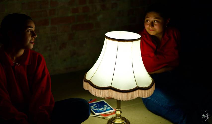 Zwei Personen in roten Hemden sitzen im Dunkeln neben einer eingeschalteten Tischlampe und einem Notizbuch auf dem Boden.