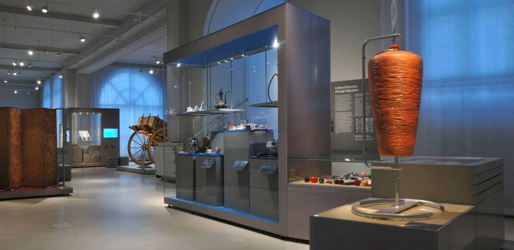 Ansicht der Dauerausstellung „Kulturkontakte. Leben in Europa“ – Modernes Museum mit beleuchteten Vitrinen, historischem Wagen und großem roten Garnspindelobjekt auf Podest. Beschriftungstafel "Cultural Diversity through