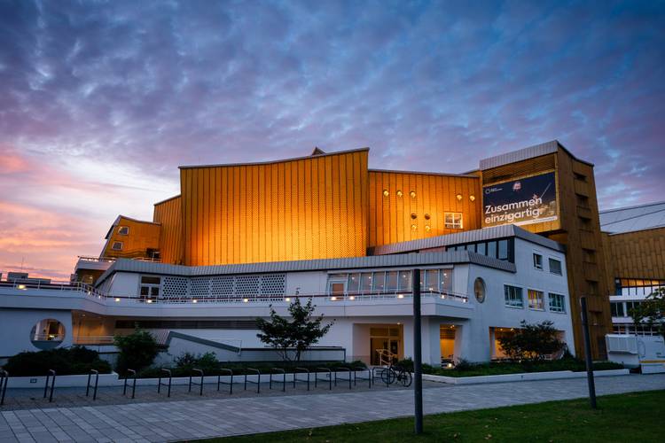 Die Philharmonie im Abendlicht – Die Philharmonie Berlin mit goldenen Panelen und weißen unteren Stockwerken wird in der Abenddämmerung beleuchtet, mit einem bewölkten Himmel im Hintergrund. Auf einem großen Banner steht auf Deutsch „Zusammen einzigartig“.
