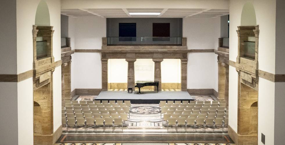 Konzerthalle mit leerem Podium, Klavier und Stuhl. Reihen leerer Stühle im Vordergrund, dekorativer Boden, Holzwände und Galerien.