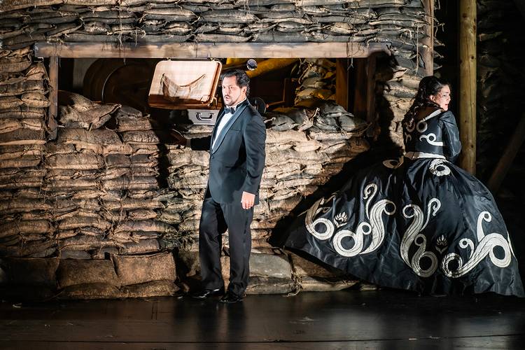 La forza del destino – Person im Anzug und person in prachtvollem, schwarz-weiß gemustertem Kleid vor einem Sandsackwall. Beide schauen in entgegengesetzte Richtungen.