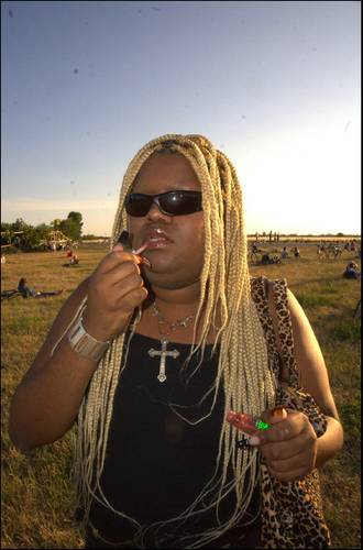 XD ERICA – Person mit blonden Zöpfen trägt Sonnenbrille und schwarze Kleidung, hält Lipgloss auf einer Wiese. Im Hintergrund Menschen und Fahrräder.