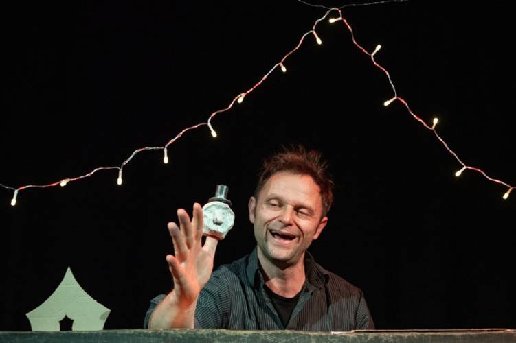 Person mit Flasche in der Hand lächelt. Im Hintergrund schwarze Wand mit Lichterkette. Unten links spitze Form sichtbar.