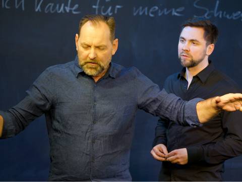  – Zwei männlich gelesene Personen vor einer Tafel. Eine Person gestikuliert. Auf der Tafel steht handschriftlich: "Ich kaufte für meine Schwester..."