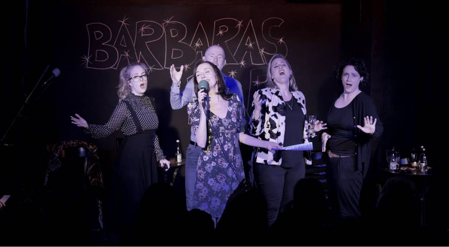 Fünf Personen singen auf einer Bühne, im Hintergrund das Schild "BARBARAS". Alle tragen dunkle Kleidung. Zwei Personen halten einen Zettel.