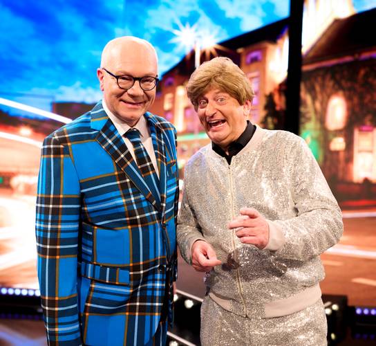 Heißmann & Rassau - LIVE 2027 – Zwei Personen, eine im blau karierten Anzug, die andere im silbernen Glitzeranzug, stehen lächelnd nebeneinander vor einer unscharfen Stadtansicht.