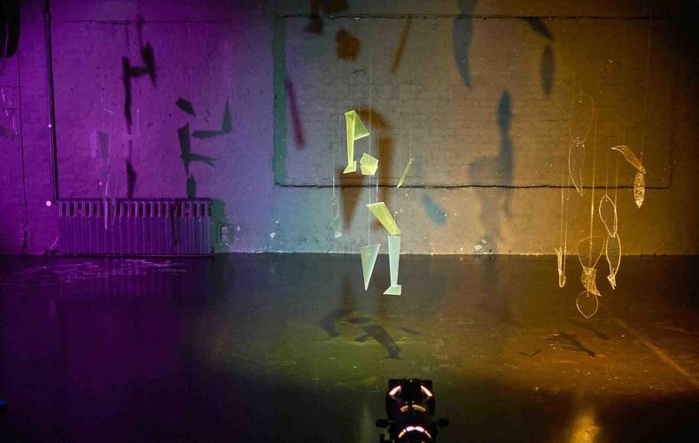 Objekte aus Draht und durchsichtigen Materialien hängen vor einer bunt beleuchteten Wand. Schattenspiele und Lichtprojektionen sind sichtbar.