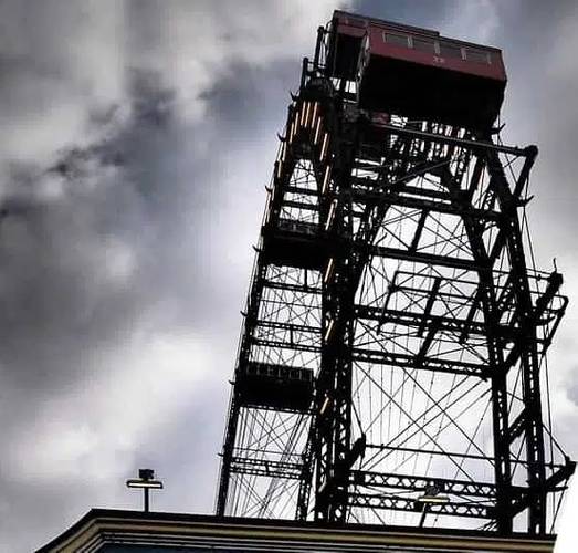 Großes Riesenrad in metallischer Struktur vor wolkigem Himmel. Oben rechts eine rote Gondel, unten links ein beleuchtetes Kreuz auf einem Dach.
