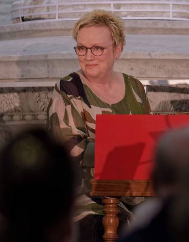Eine Person mit kurzen blonden Haaren und Brille sitzt an einem Tisch mit rotem Aufsteller, im Hintergrund Marmordekor.