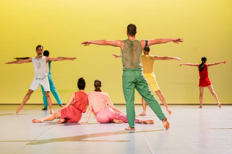 20 Jahre Radialsystem: In C - special performance 1 – Sieben Tänzer*innen in bunten Klamotten in Bewegung auf einer Bühne vor gelben Hintergrund.