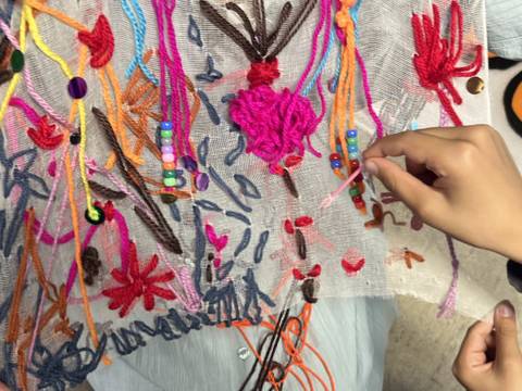  – Zwei Hände sticken bunte Muster aus Garn und Perlen auf transparentem Stoff. Person trägt blaugraues Outfit. Stickerei mit abstrakten Formen.