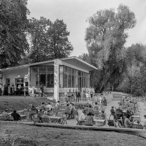 Milchhäuschen am Weißensee von Ludmilla Herzenstein, um 1978, Berlin-Weißensee, Parkstraße 33a – Schwarz-Weiß-Fotografie des Milchhäuschens in Berlin-Weißensee, vor dem viele Menschen in kleinen Grüppchen an Tischen im Außenbereich sitzen. Das Milchhäuschen wird von hohen Bäumen gesäumt.