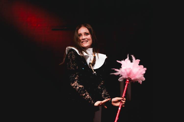 Foto: Michał Wojtarowicz – Person in schwarzem Spitzenkostüm mit großem weißen Kragen hält einen pinken Federwedel, dunkler Hintergrund mit rotem Licht.