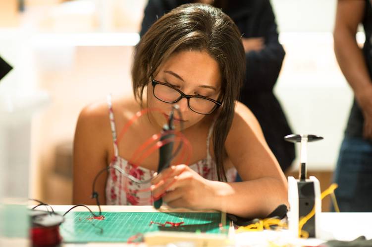Mit recylebarem Plastik fantastische Dinge in 3D zeichnen. – Ein Mädchen sitzt an einem Tisch vor einer grünen Bastelunterlage. Sie hält einen schwarzen 3D-Stift in ihrer linken Hand. Mit dem im Stift erwärmten, roten Plastik zeichnet das Mädchen etwas auf die Unterlage.