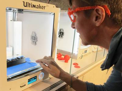  – Man sieht ein Foto, das den 3D-Drucker "Ultimaker" zeigt. Dazu ein junger Mensch, der mit seiner Hand den Drucker bedient.