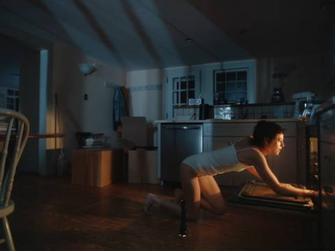 Olivia Erlanger, Appliance, Filmstill, 2024 – Person im Nachthemd kniet in dunkler Küche vor offenem Ofen. Kartons und Tisch im Hintergrund. Stimmungsvolle Beleuchtung. Filmstill.