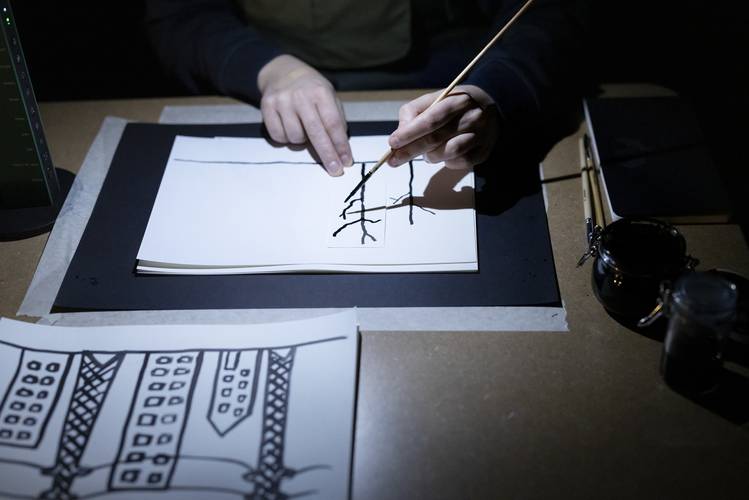 Plateau – Person zeichnet mit Pinsel schwarze Linien auf Papier; daneben Tinte. Im Vordergrund Zeichnung von Hochhäusern.