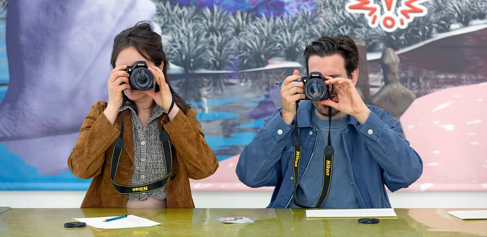 Zwei Personen fotografieren mit Nikon Kameras. Im Hintergrund ein farbenfrohes Gemälde mit Ausrufezeichen. Auf dem Tisch liegen Papier und Stifte.